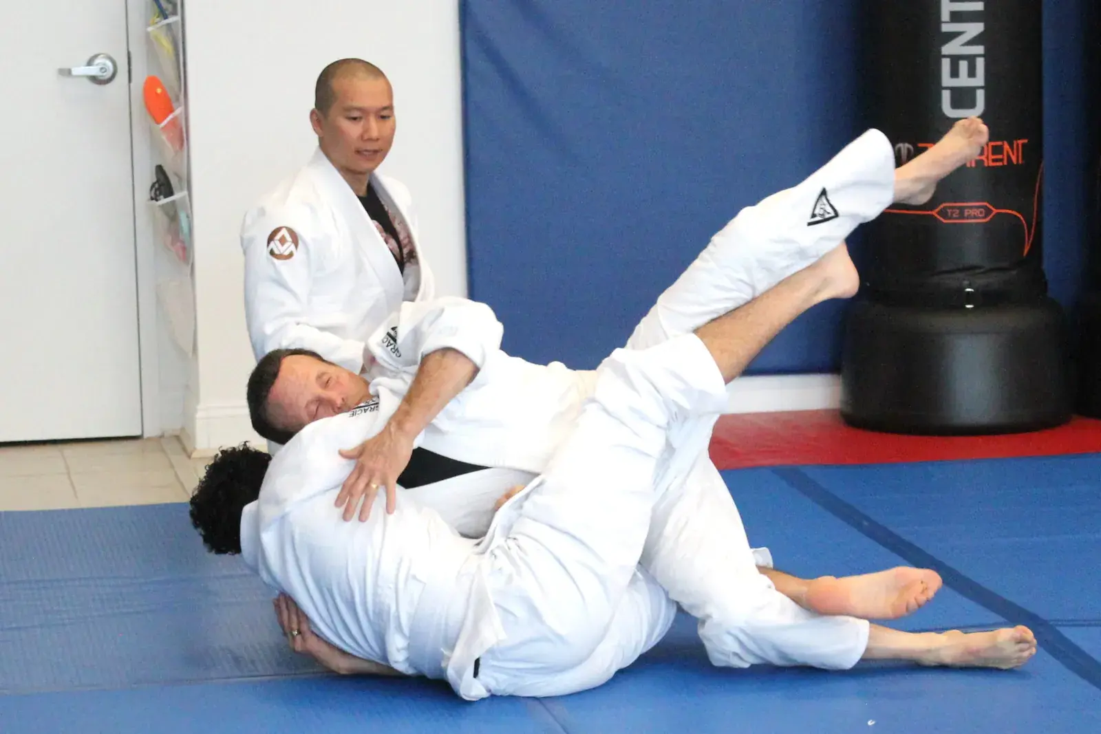 Adults practicing a jiu-jitsu sweep as an instructor watches