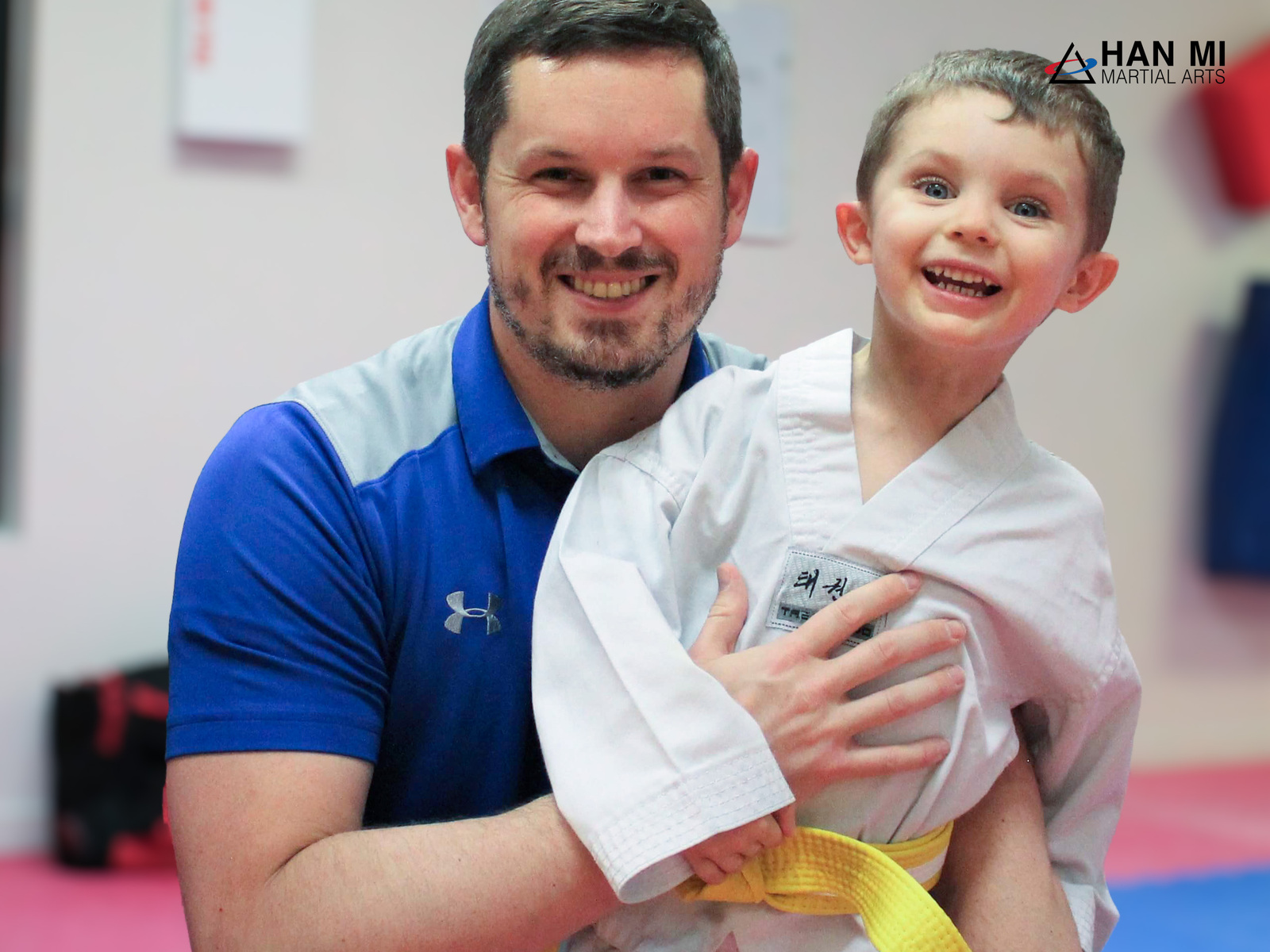 A Han Mi coach holding a smiling Little Dragons student with a yellow belt