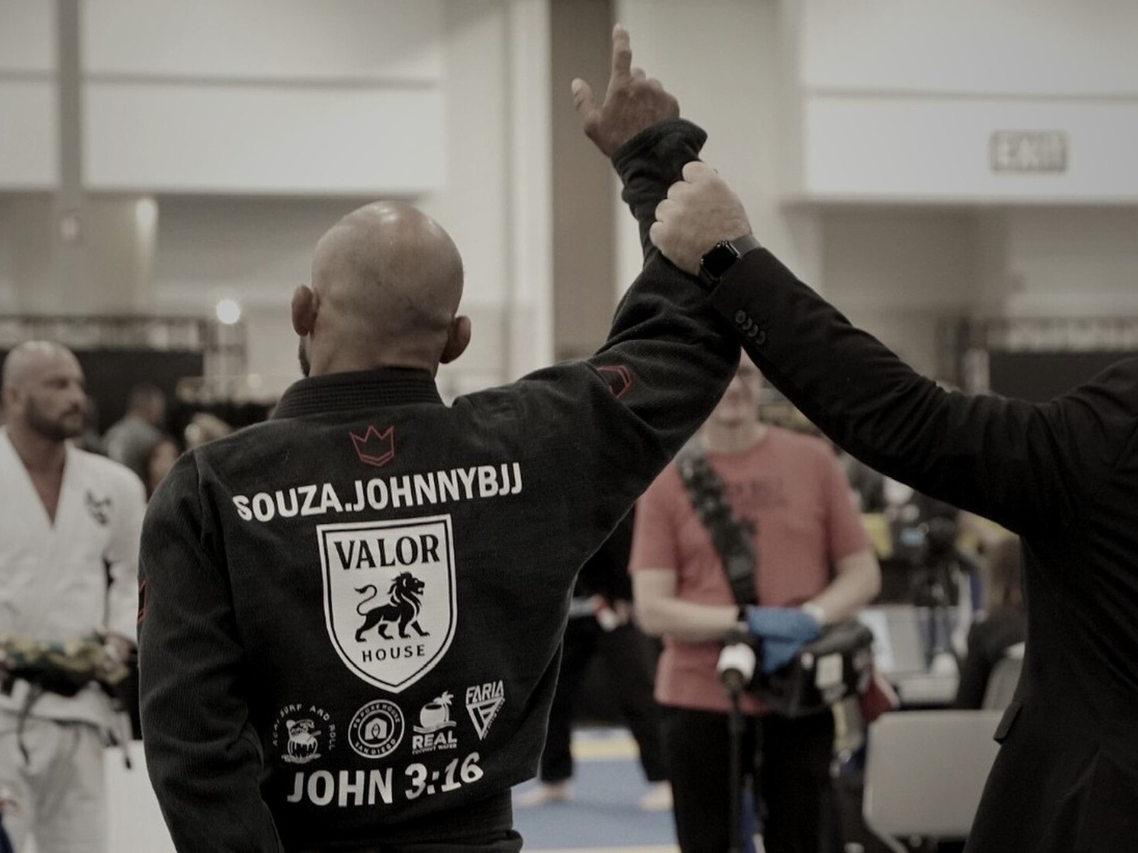 Johnny seen from behind at a competition, his arm raised and the Valor House crest visible on his gi.