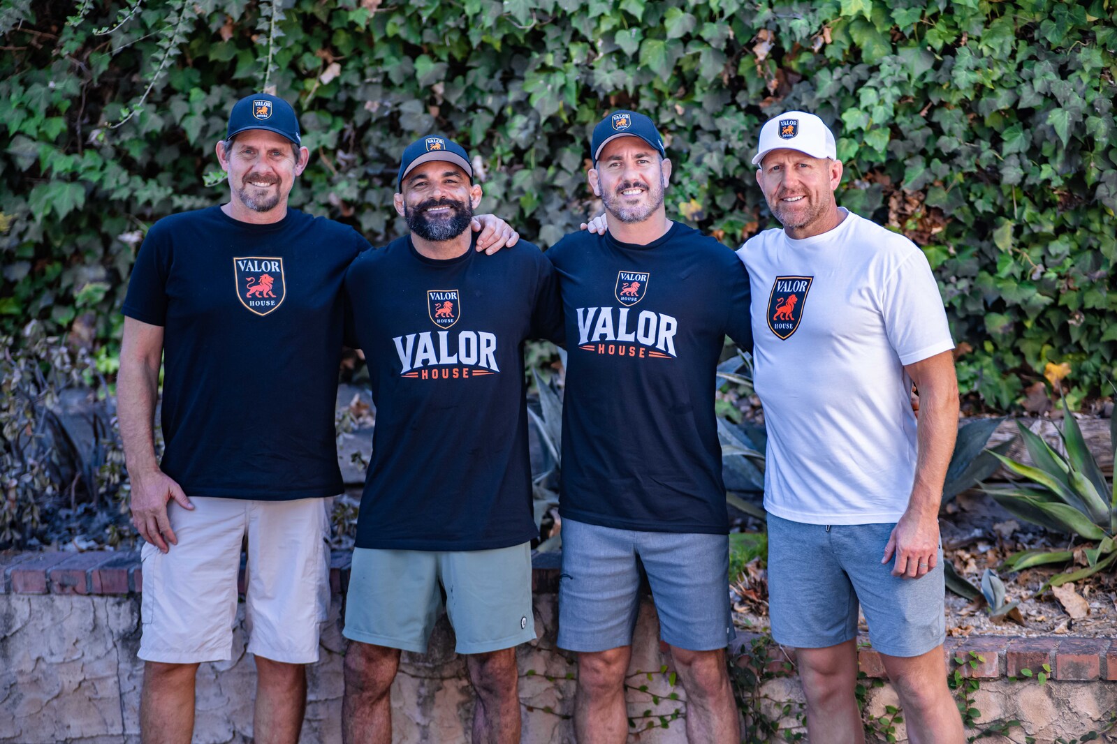 Four people wearing Valor House apparel, standing together against an ivy-covered wall.