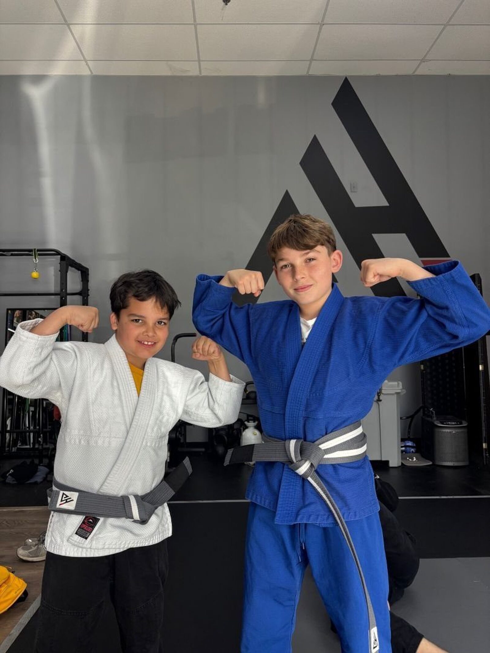 Two kids in gis flexing in front of the academy logo