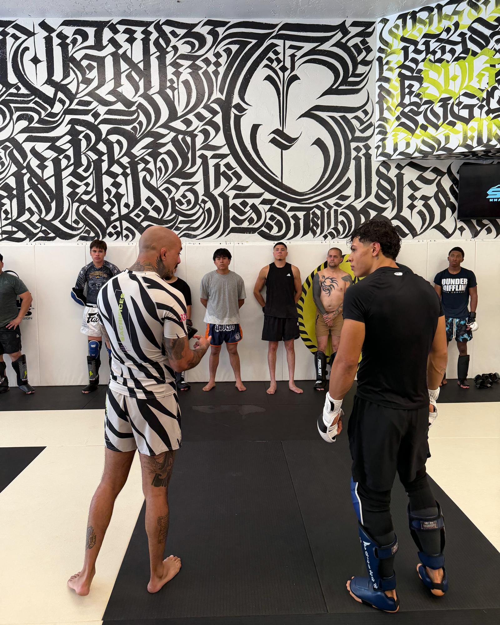 SVG coach in a zebra-stripe rashguard standing face to face with a fighter in shin guards while the class lines the wall behind them, under the black-and-white calligraphy mural