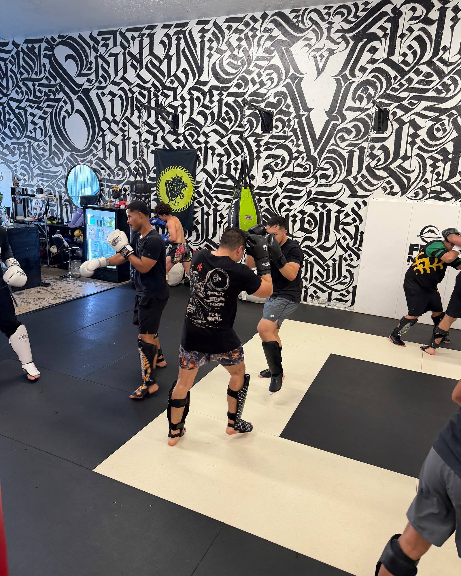 SVG members in gloves and shin guards drilling striking in pairs on the black-and-white mats, with the calligraphy mural and lime SVG banner behind them