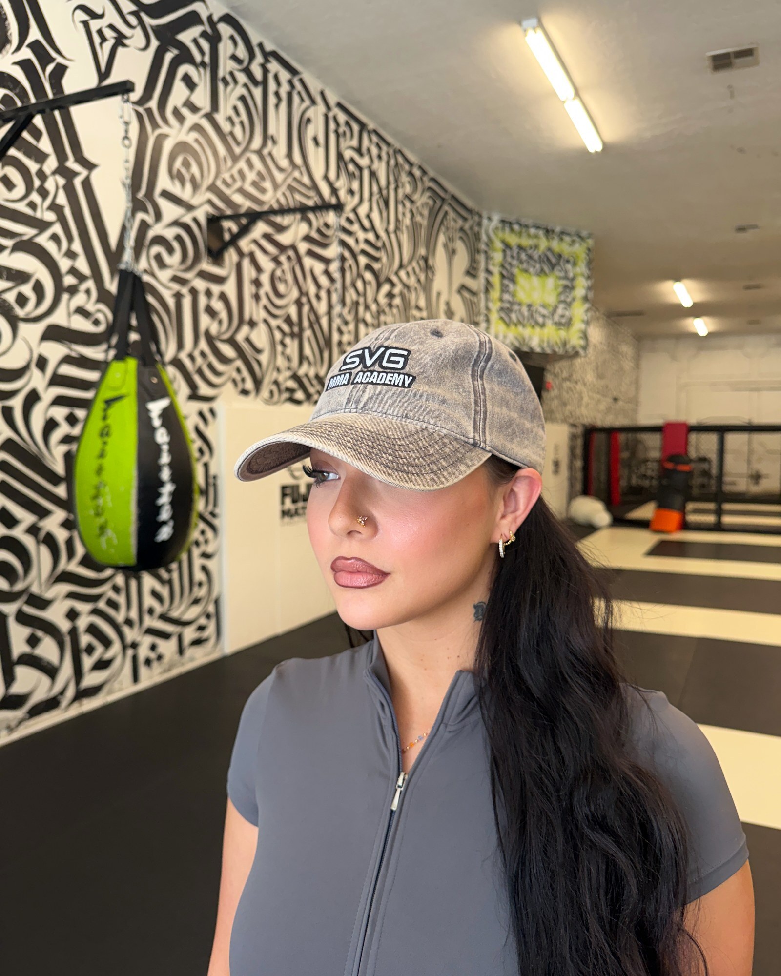 SVG member wearing a washed SVG MMA Academy cap inside the gym, with the calligraphy mural and a lime hanging bag behind her