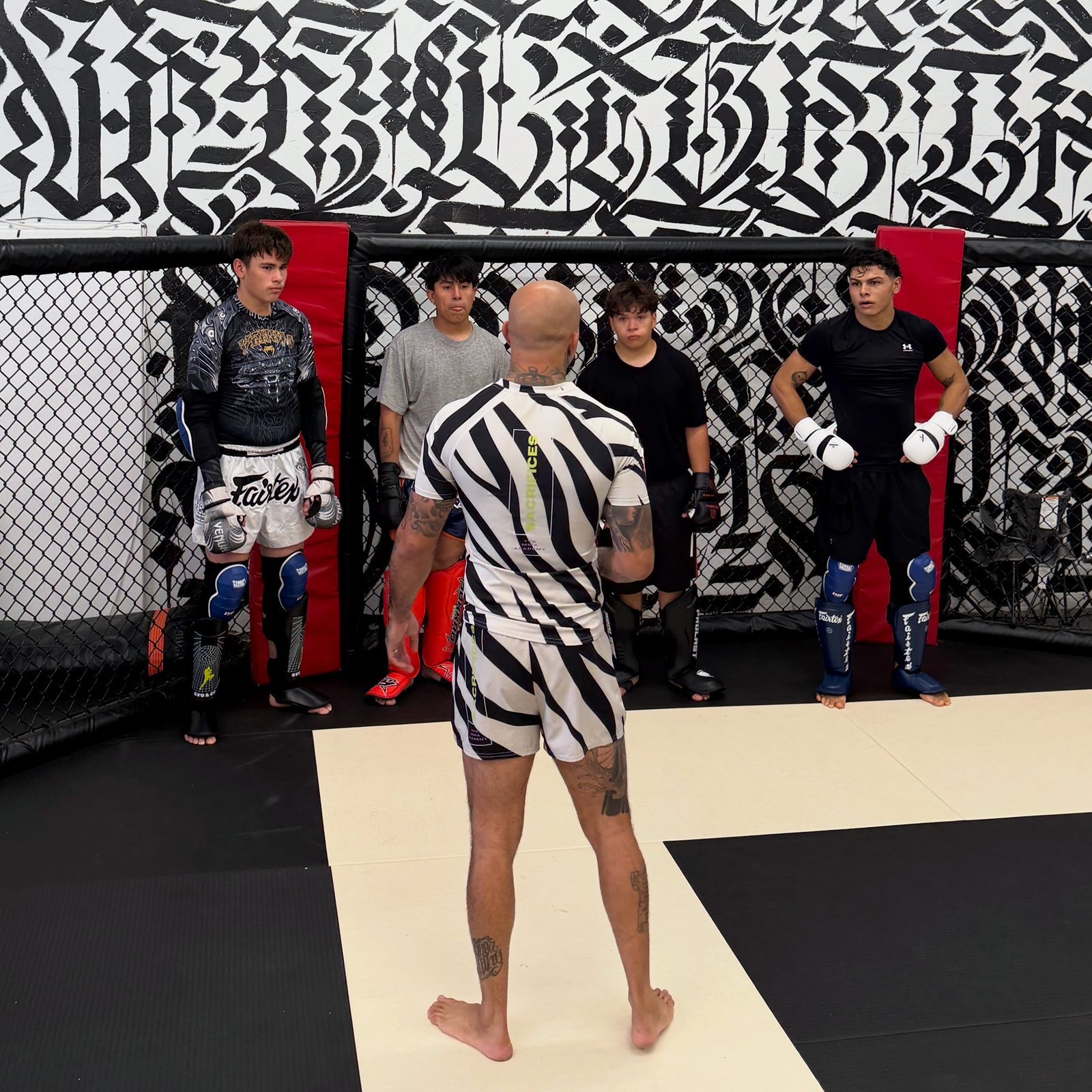 SVG coach in a zebra-stripe rashguard talking four students in gloves and shin guards through the plan inside the SVG cage