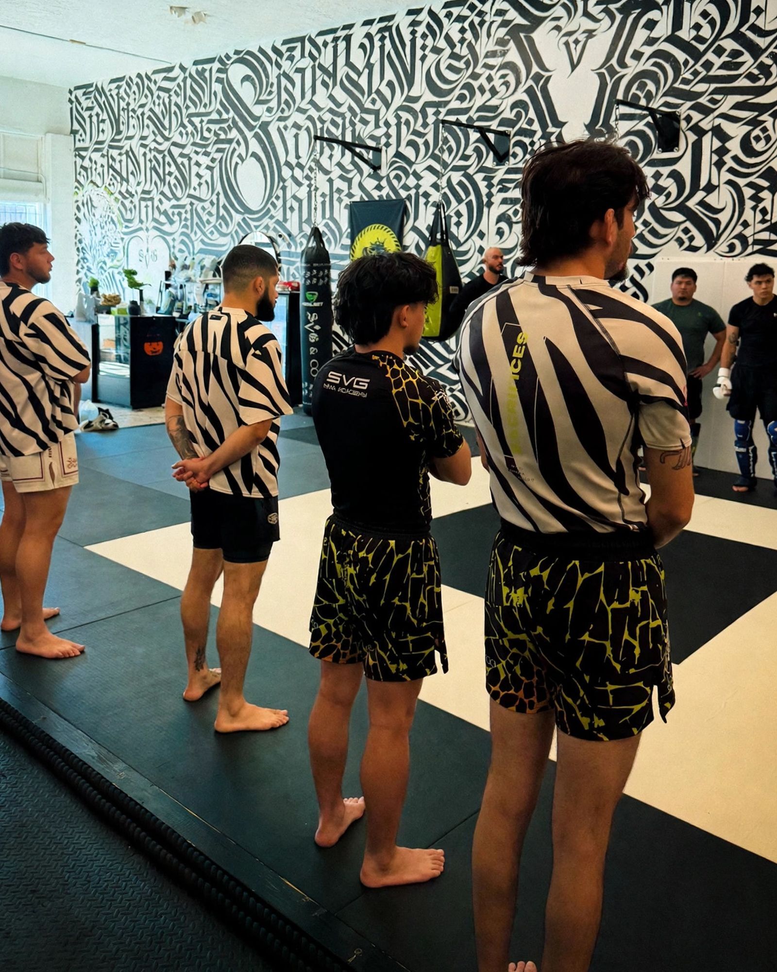 SVG team members in zebra-stripe and lime-print fight shorts lined up barefoot on the mats to greet a new member, facing the calligraphy mural